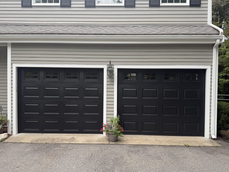 Close view of two new black raised-panel garage doors with a row of windows on a gray colonial