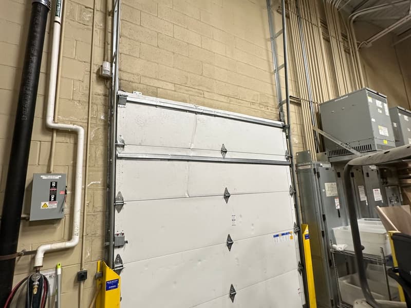 Tall white sectional door in a warehouse, with an electrical disconnect box on the block wall beside it