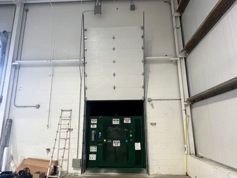 Tall commercial sectional door raised partway in a warehouse, a stepladder beside it